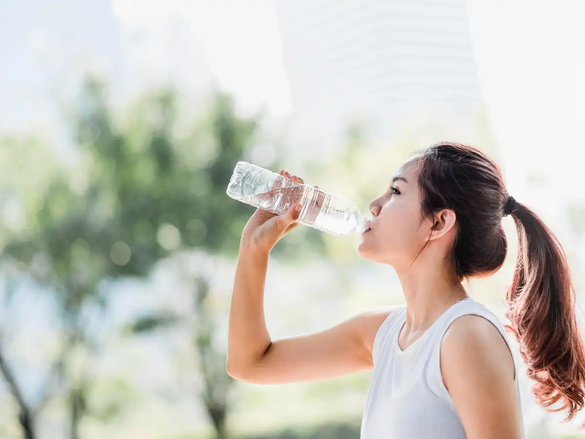 木々とビルがぼんやり写る背景。一本結びの女性がペットボトルから水を飲む画像